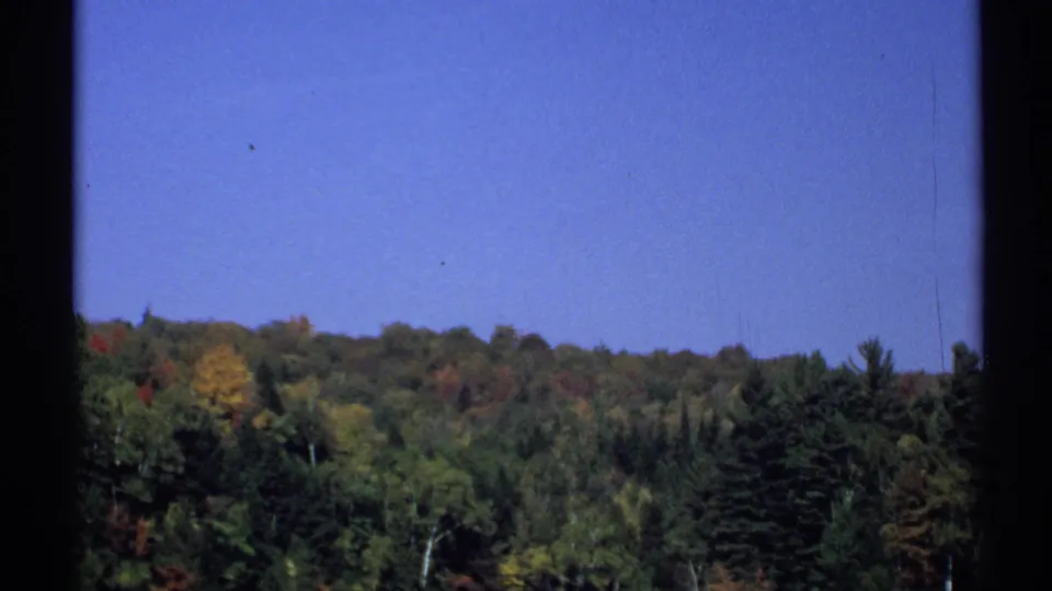 1971 New York 8mm frame showing a distant tree-covered ridge with green, yellow, and red autumn foliage under a clear blue sky.