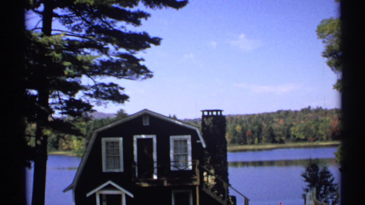 1971 New York 8mm frame of a dark cabin with white trim and stone chimney beside a lake under clear sky.