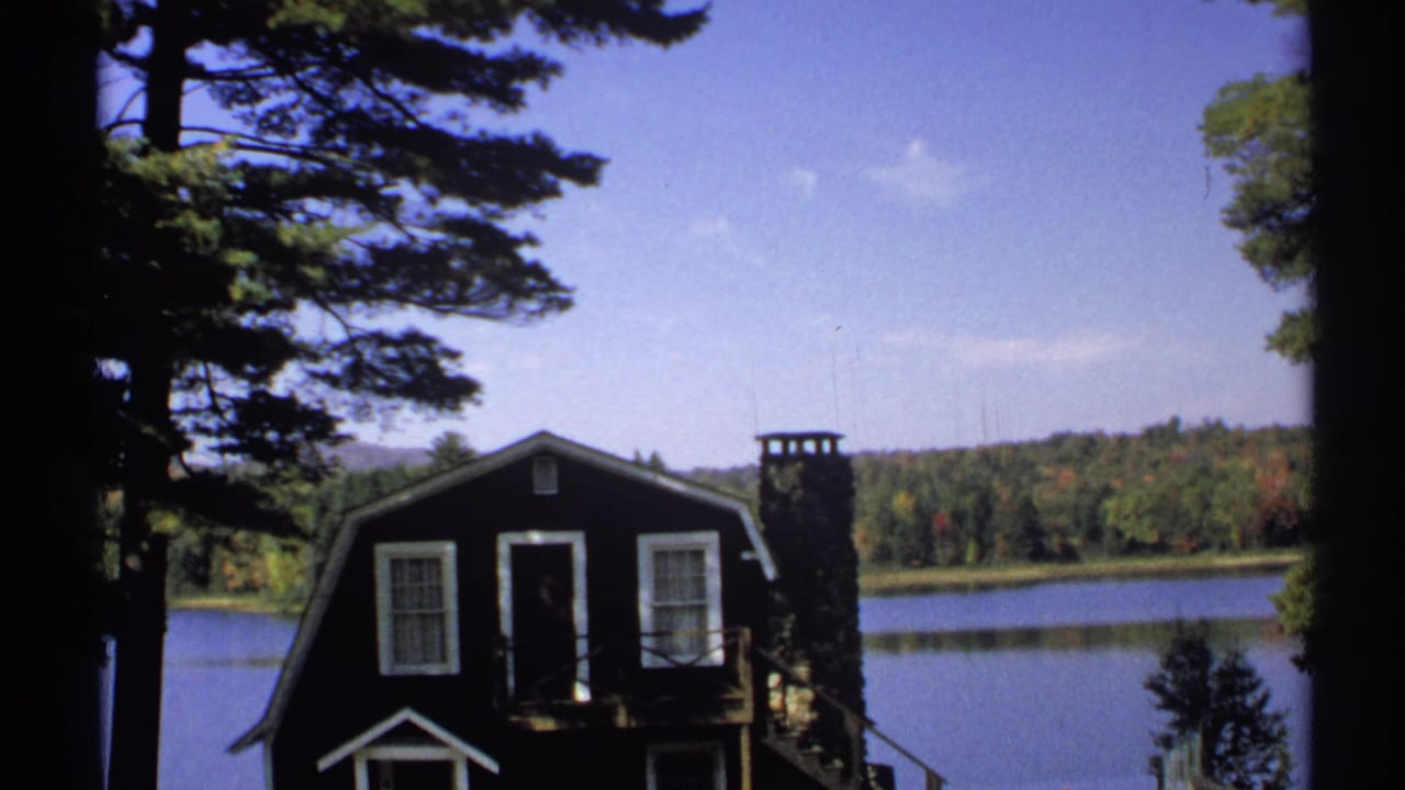 1971 New York home-movie frame showing a dark cabin, pine trees, lake water, and distant wooded hills.
