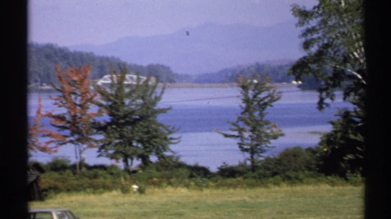 1971 New York lakeside view with trees in the foreground, calm water, distant hills, and a possible bridge across the lake.