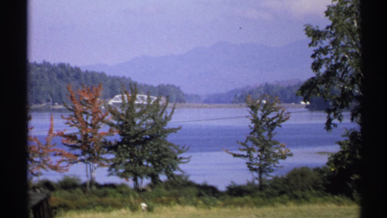 1971 New York home-movie view across a grassy lakeshore toward trees, water, wooded hills, and hazy mountains.