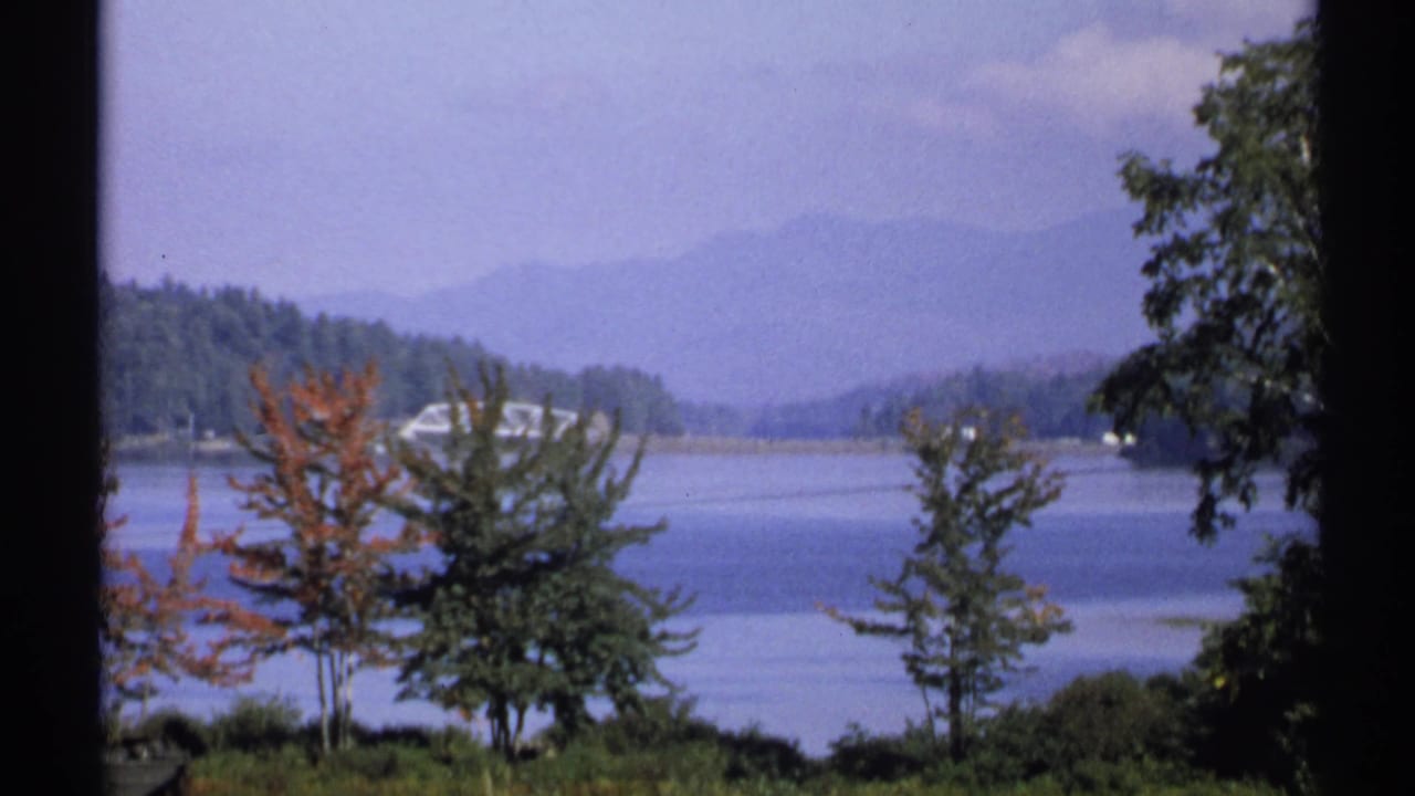 1971 New York home-movie frame showing a quiet lake, foreground trees, wooded shoreline, and blue-gray mountains.