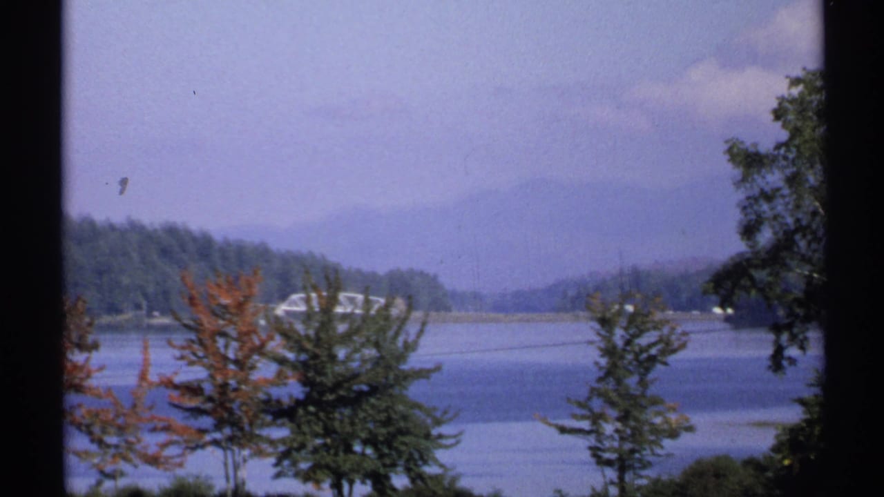 1971 New York 8mm frame showing a calm lake, autumn trees, and distant wooded hills beneath a hazy sky.