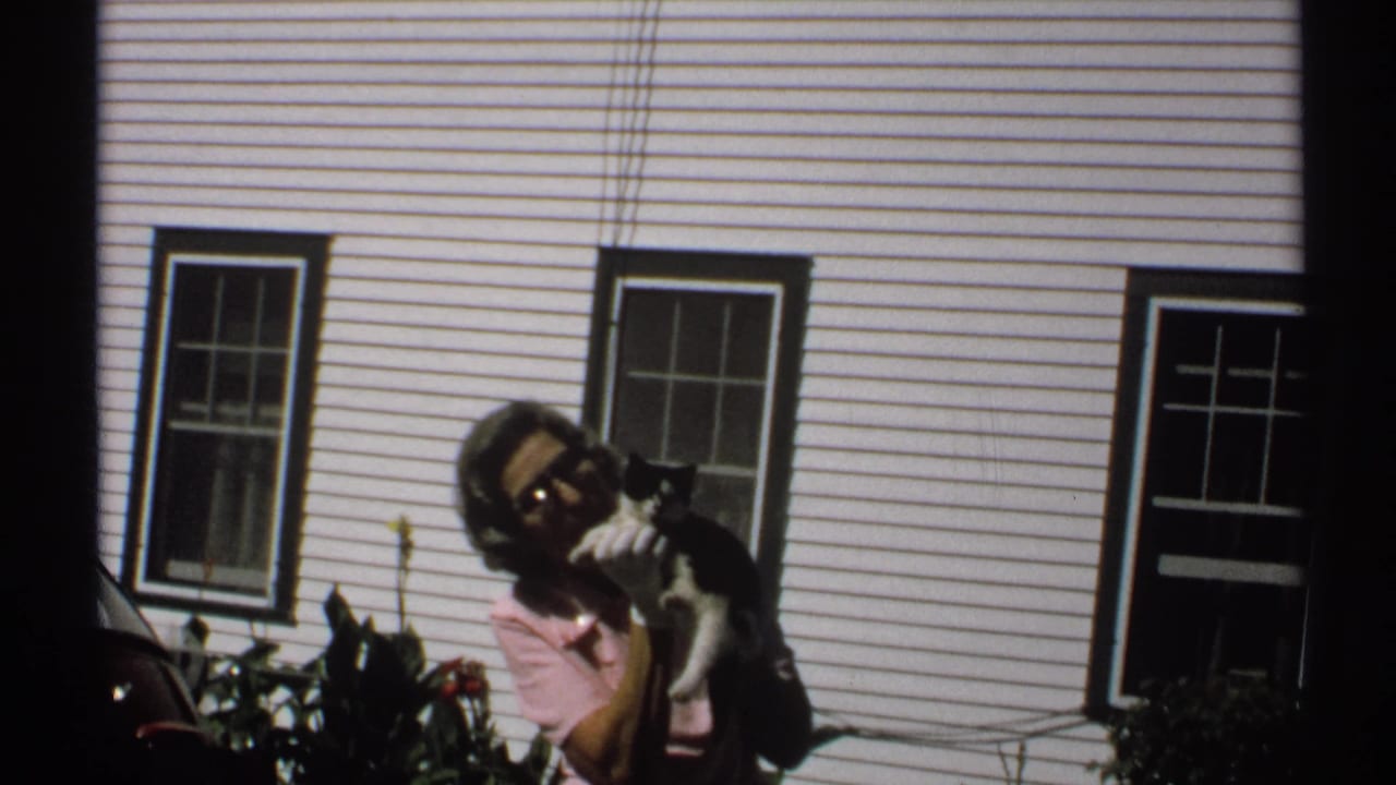 1971 New York home-movie frame with a woman facing the camera, holding a black-and-white cat beside a white paneled house.