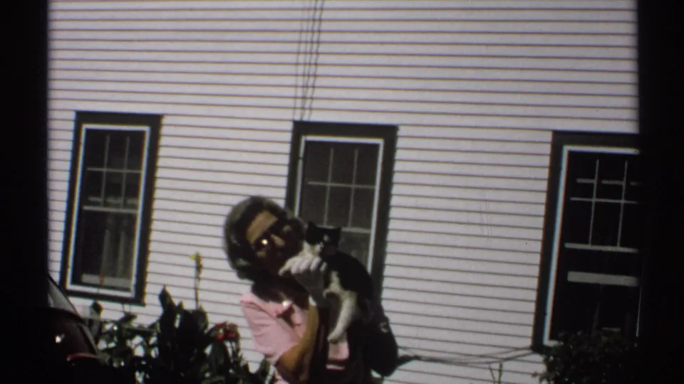 1971 New York home-movie frame with a woman facing the camera, holding a black-and-white cat beside a white paneled house.