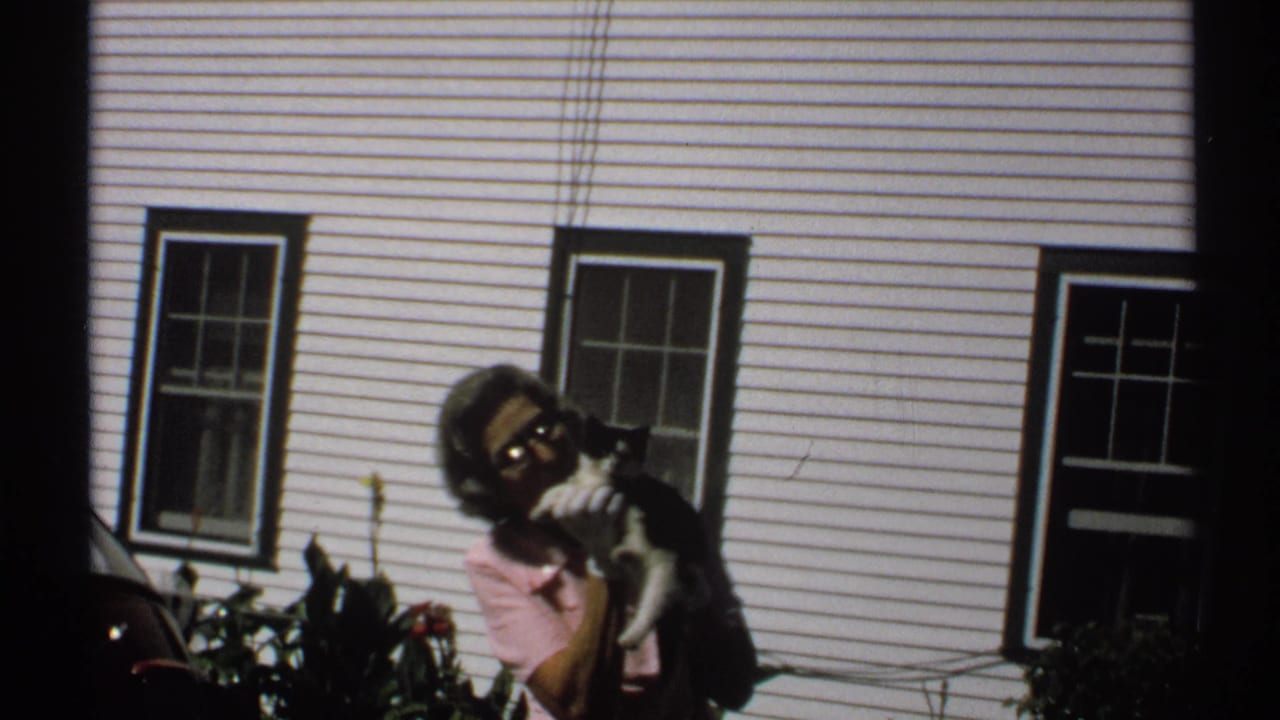 In 1971 New York home-movie footage, a woman in a pink shirt holds a black and white cat near a white clapboard building.