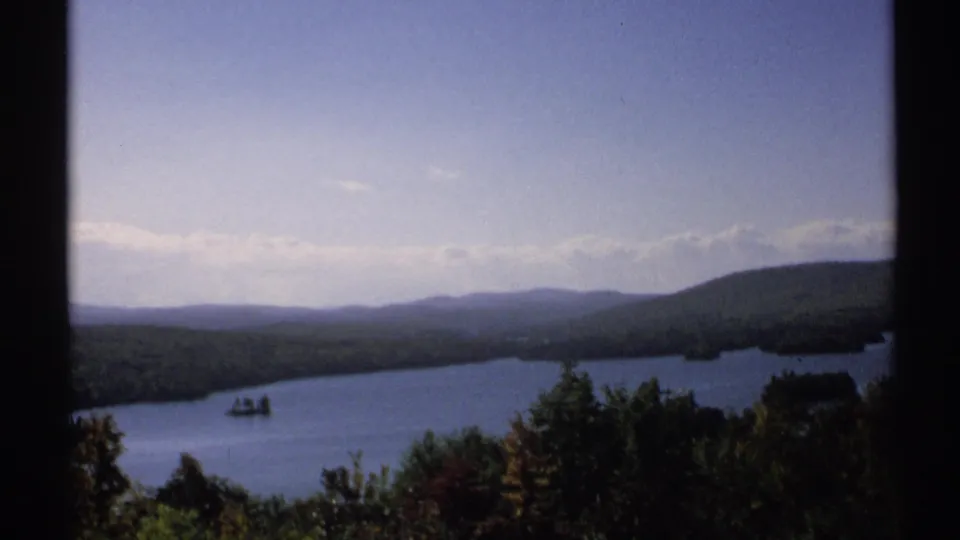 1971 New York home-movie view of a lake, wooded shoreline, distant hills, and pale sky divided by a dark frame.