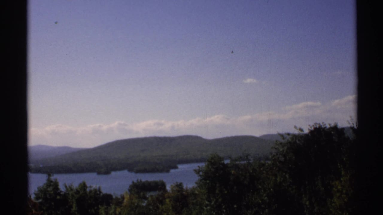 1971 New York home-movie frame of a lake below rolling forested hills, with foreground trees and dark frame borders.