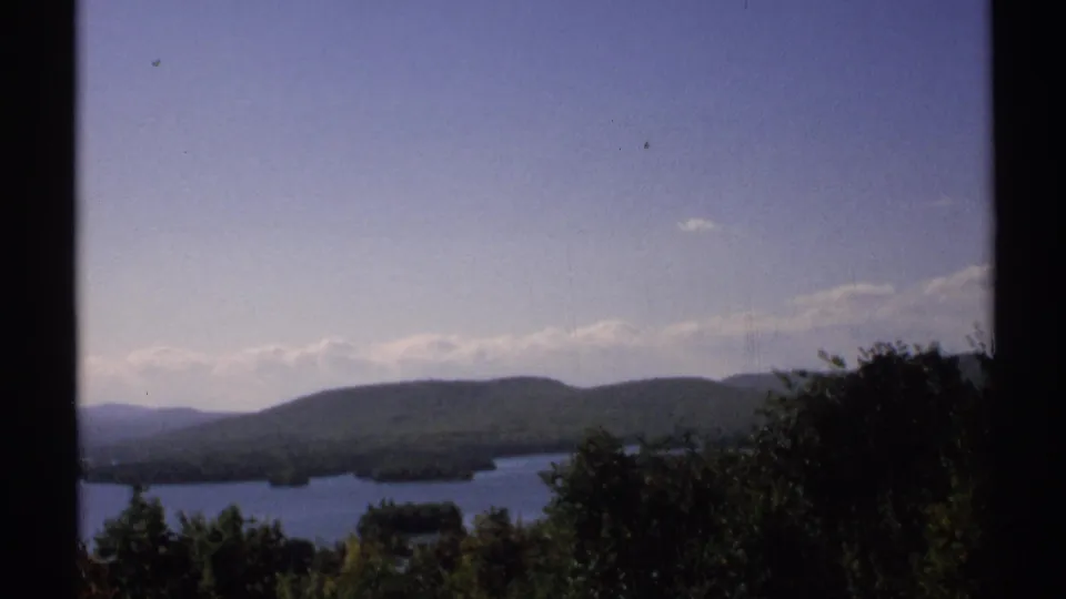 1971 New York home-movie frame of a lake below rolling forested hills, with foreground trees and dark frame borders.