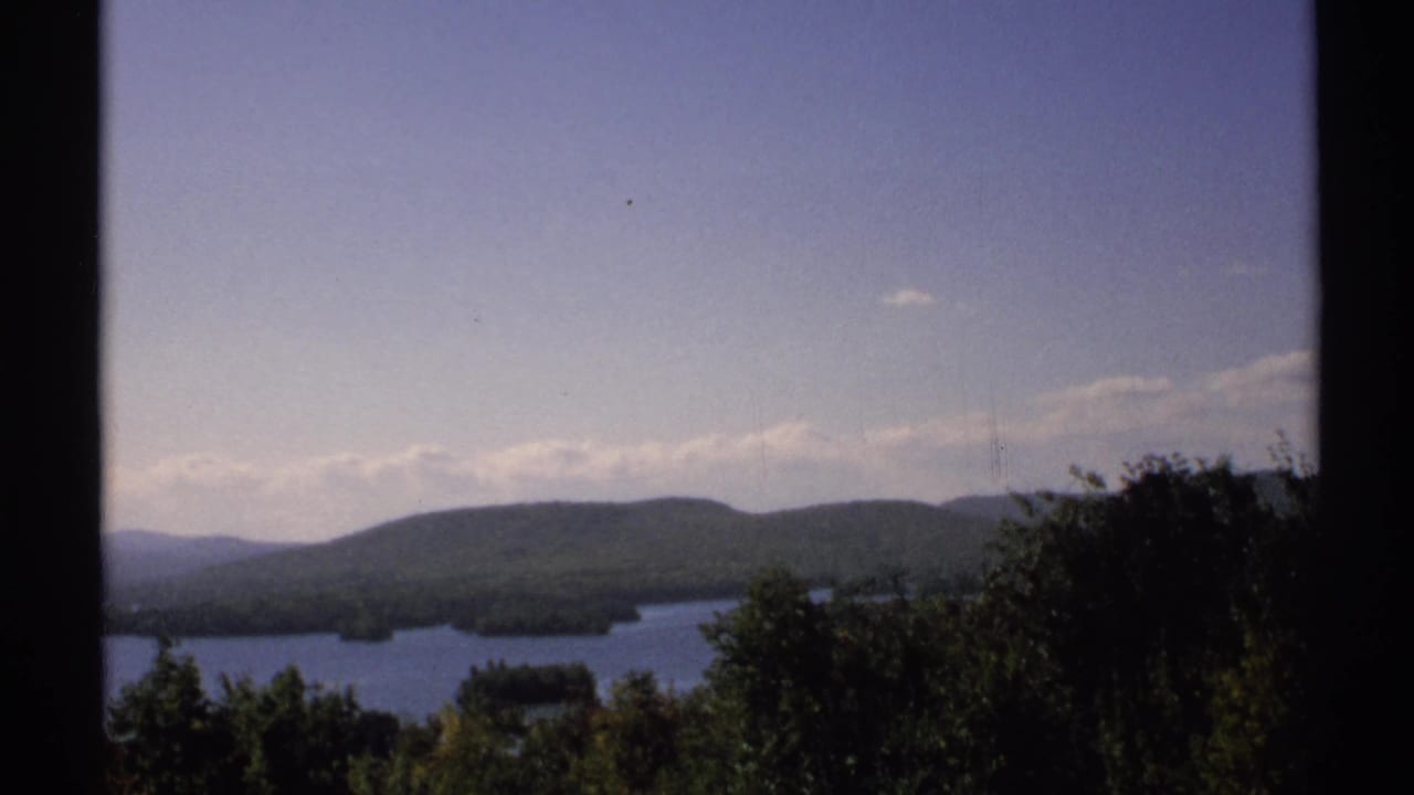 1971 New York scenic lake view with dense foreground trees, distant wooded hills, small islands, and a dark vertical edge.