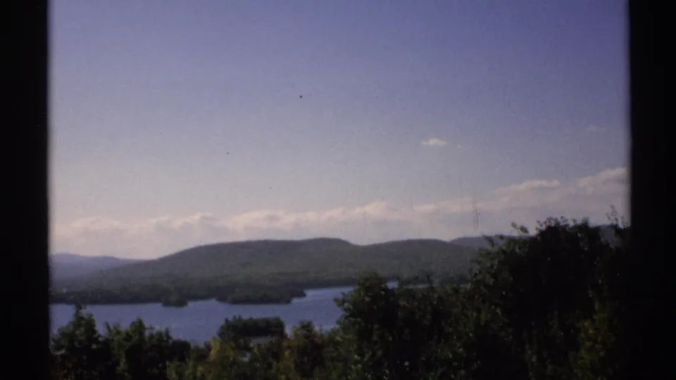 1971 New York scenic lake view with dense foreground trees, distant wooded hills, small islands, and a dark vertical edge.