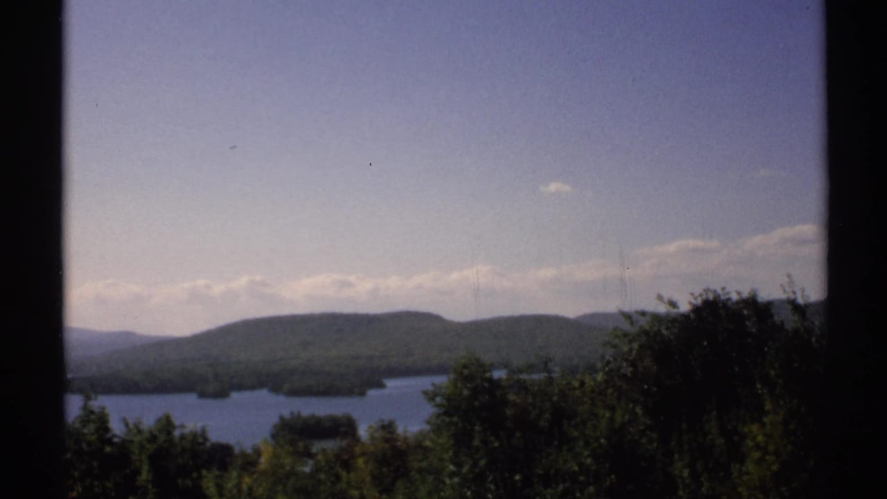1971 New York 8mm frame showing a calm lake beyond trees with low forested hills and pale clouds in the distance.