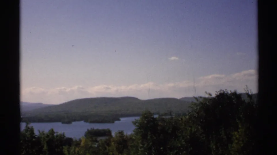 1971 New York 8mm frame showing a calm lake beyond trees with low forested hills and pale clouds in the distance.