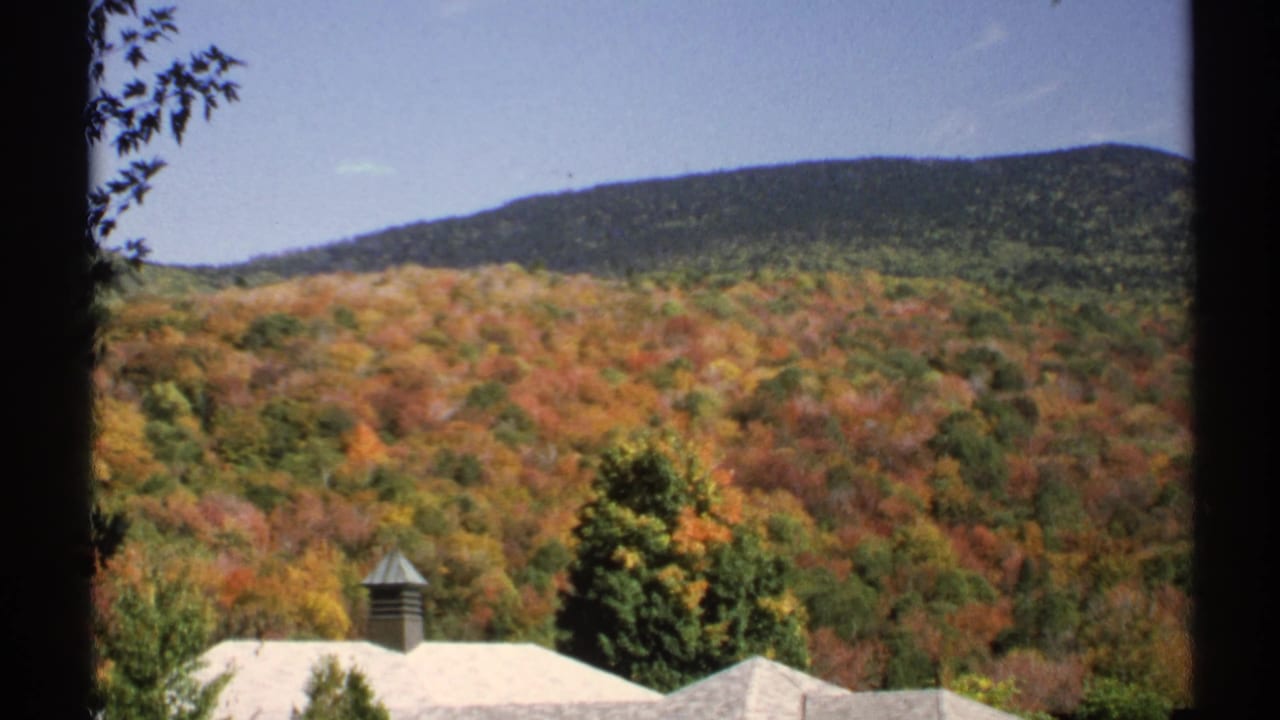 1971 New York 8mm frame holding on forested fall hills with a pale-roofed house and chimney partly visible in the foreground.