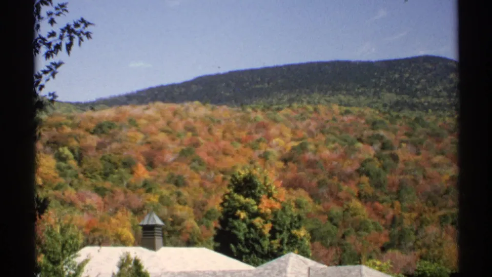 1971 New York 8mm frame holding on forested fall hills with a pale-roofed house and chimney partly visible in the foreground.