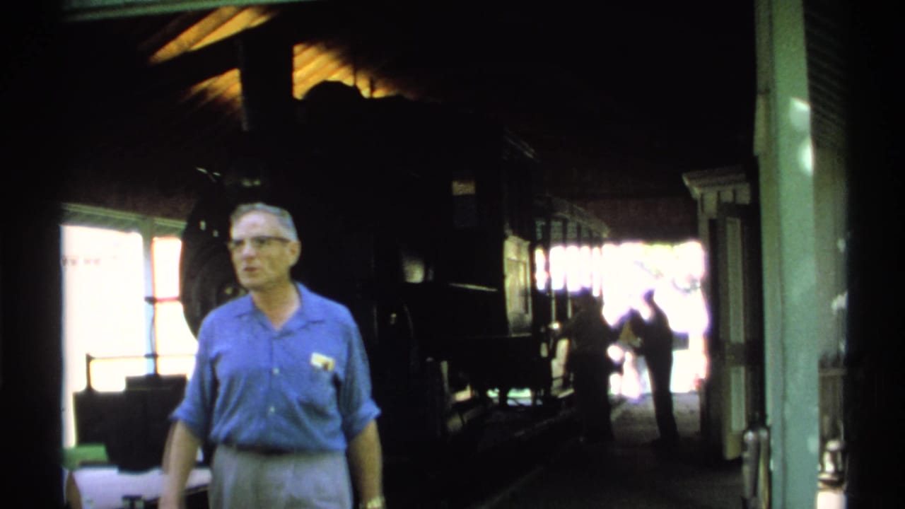 1971 New York 8mm frame showing a man in a blue shirt near a shadowed steam locomotive and bright station entrance.