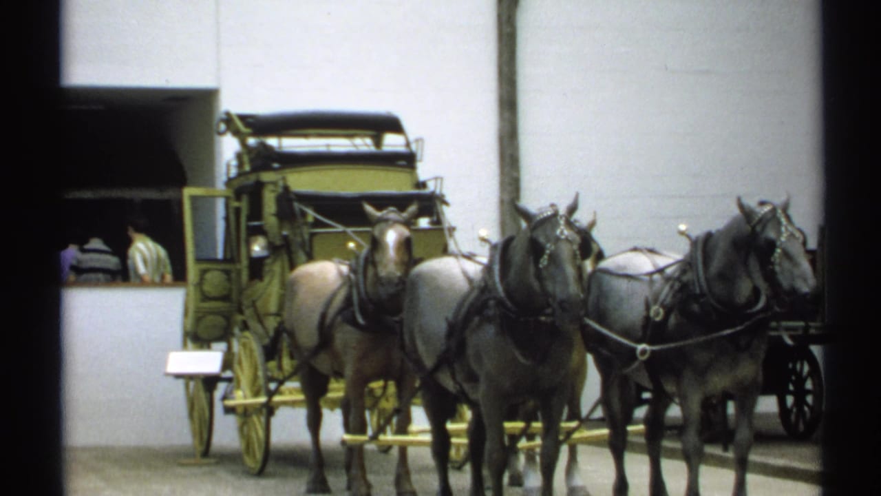 Indoor museum display with harnessed horses, a carriage, and visitors near a doorway.