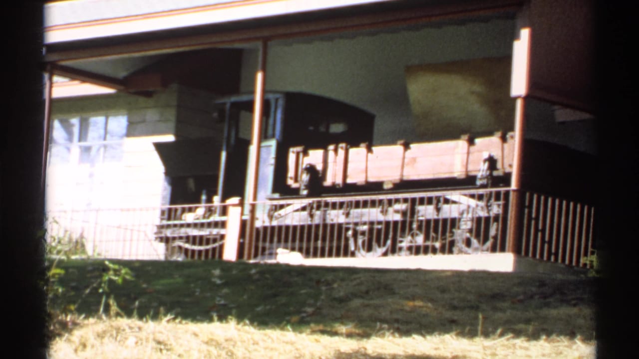 1971 New York home-movie frame of a wooden rail car exhibit seen across grass and a metal railing, with small visitors near the car.