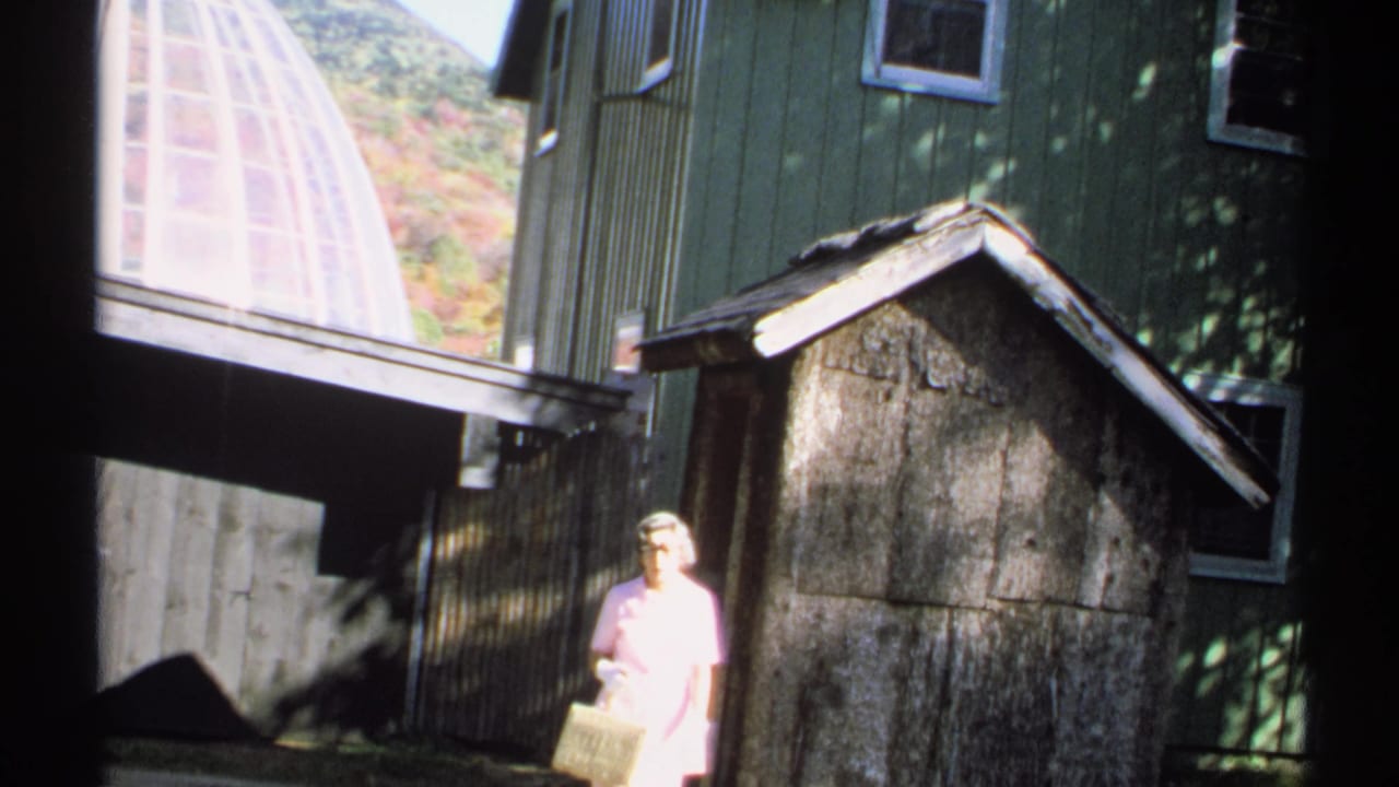 1971 New York home-movie frame showing a woman in pink with a basket beside a rustic shed and green house, with hills visible beyond.