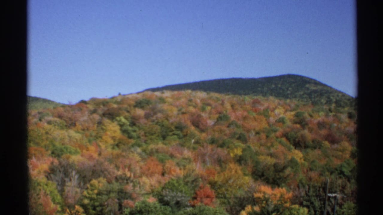 1971 New York 8mm frame showing red, orange, and green foliage on a hillside with a distant mountain ridge under clear blue sky.