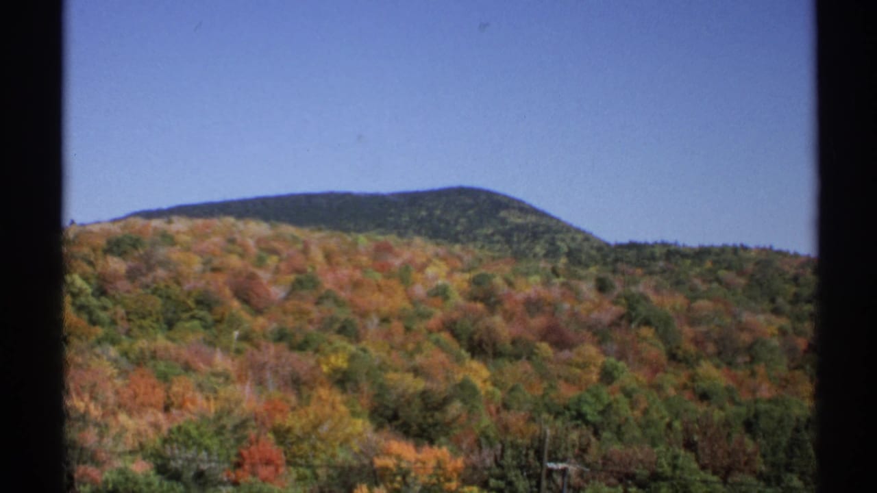 1971 home-movie frame from New York showing a broad autumn hillside, mixed red and orange trees, and a low ridge against blue sky.