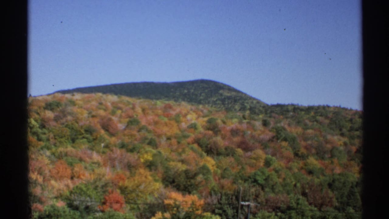 8mm New York landscape frame with red, orange, and green autumn woods across a hillside and a dark ridge line in the background.