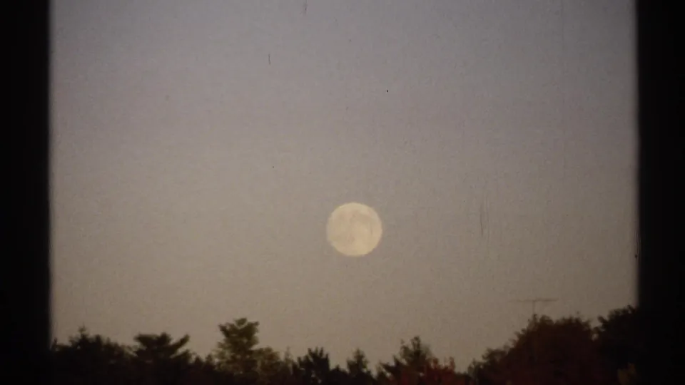 1971 New York home-movie frame showing a pale sun over a dark shoreline or treeline near water.