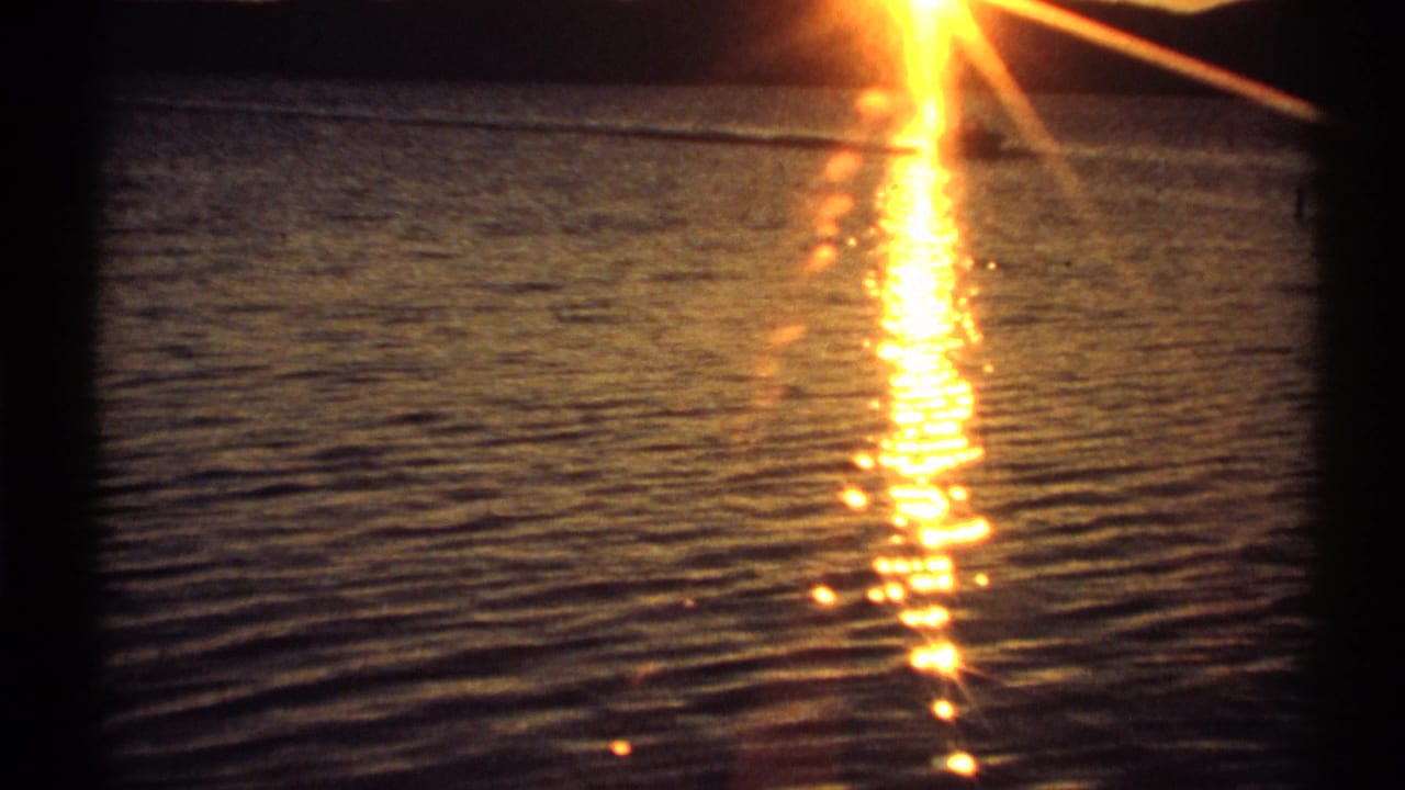 Vintage 8mm New York waterfront view with bright sunset glare mirrored in dark, textured water.