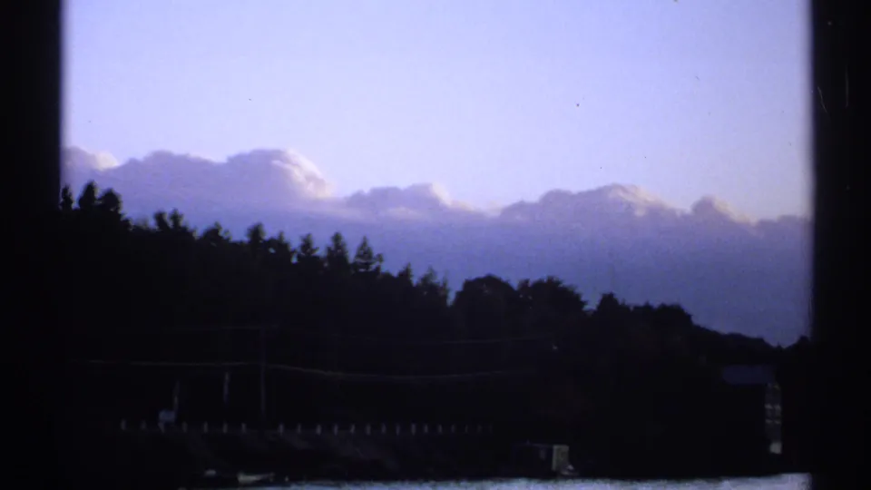1971 New York home-movie frame of a wooded shoreline under clouds, with window framing and a dim waterside structure visible below.