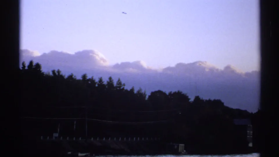 1971 New York 8mm frame showing a silhouetted treeline and low hill under layered clouds and pale blue sky.