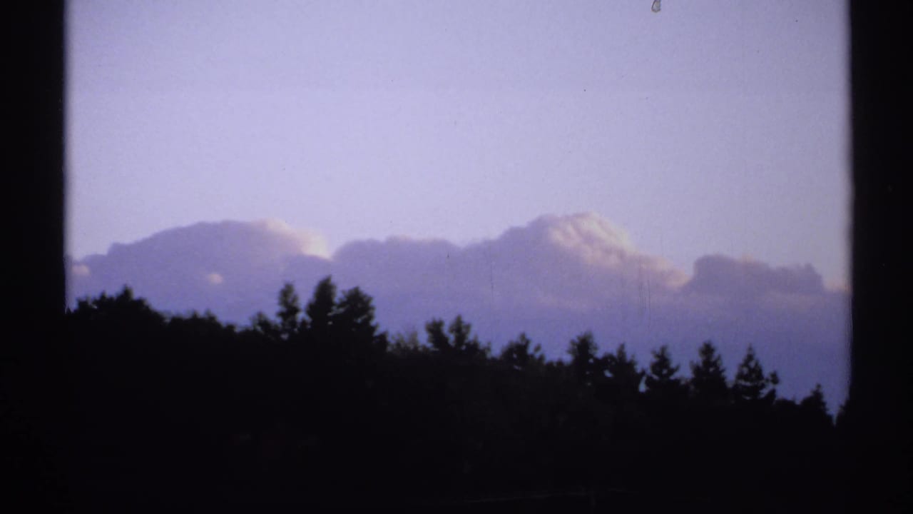 1971 New York 8mm frame showing a dark tree-covered ridge beneath pale sky and layered cloud shapes.