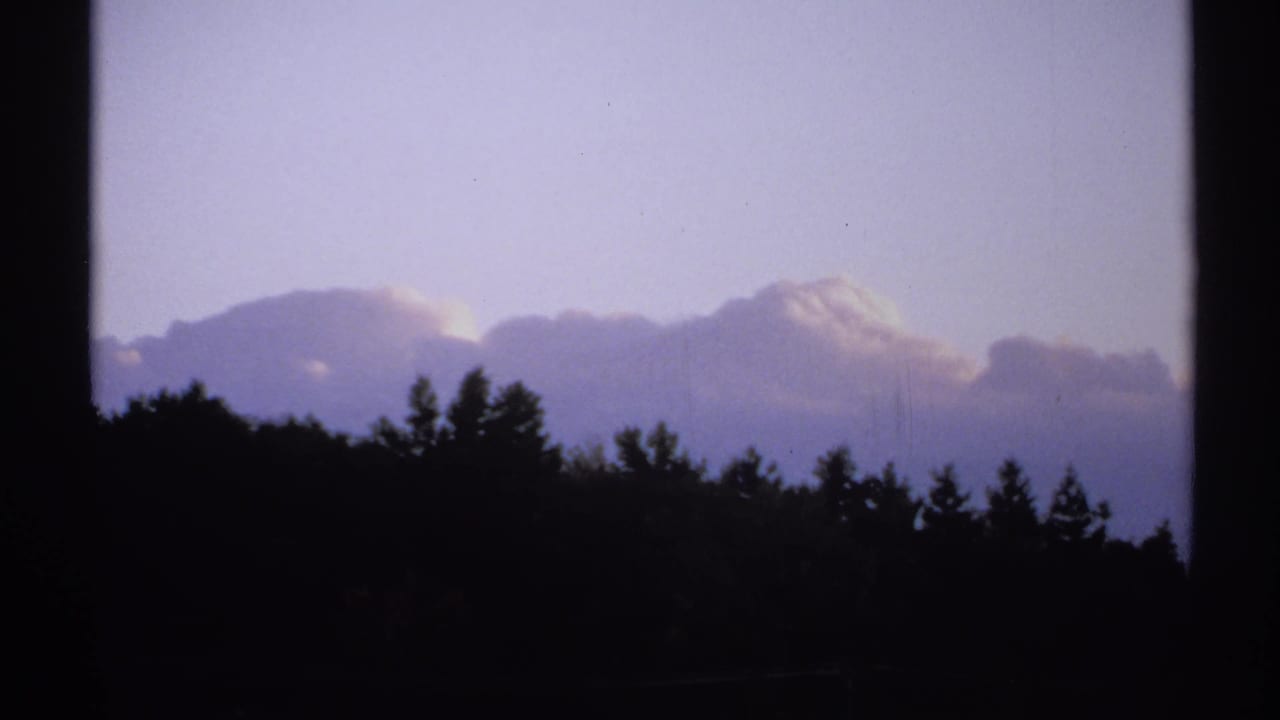 1971 New York 8mm frame with silhouetted treetops and hillside beneath broad clouds in a pale blue sky.