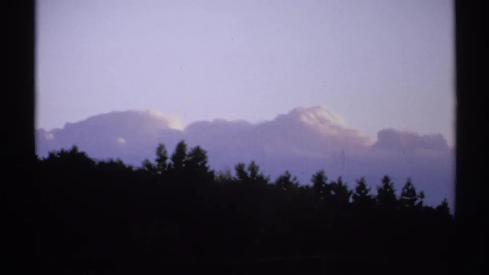 1971 New York 8mm frame with silhouetted treetops and hillside beneath broad clouds in a pale blue sky.