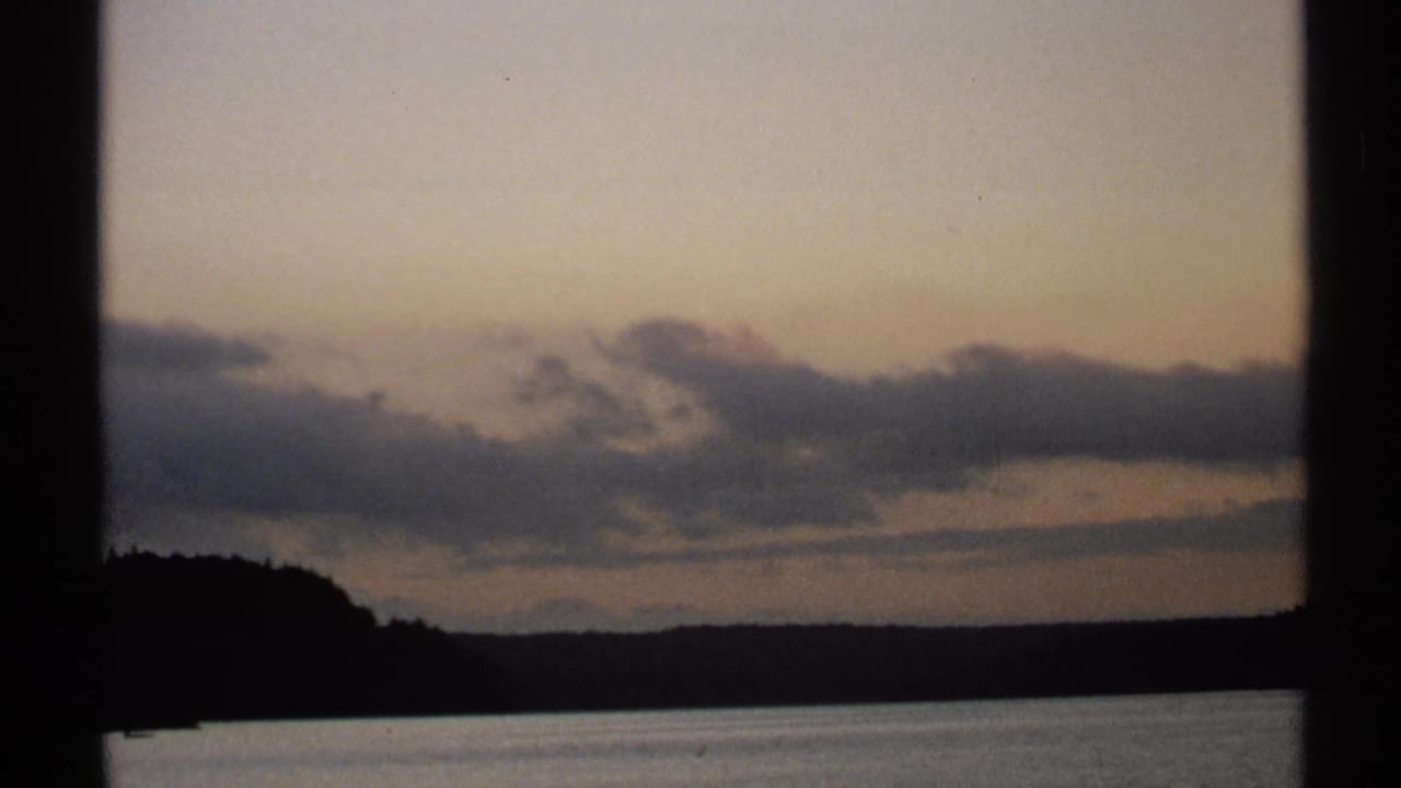 1971 New York 8mm frame showing layered dusk clouds above calm water and a silhouetted tree-lined shore.