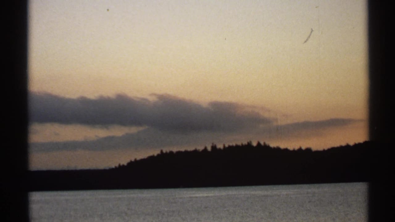 1971 New York 8mm frame showing a dark tree ridge, calm water, and warm evening clouds over the horizon.