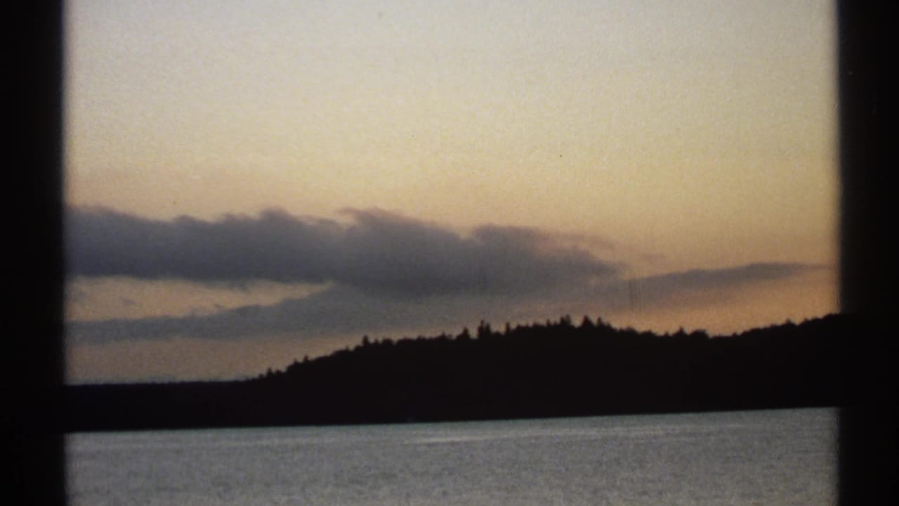 1971 New York 8mm frame with a cloud band above silhouetted trees and a smooth water surface at dusk.