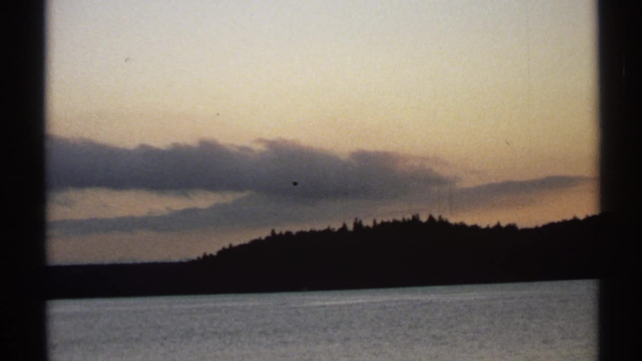 1971 New York 8mm frame of a dark lakeside shoreline, still water, and layered clouds under a fading sunset sky.
