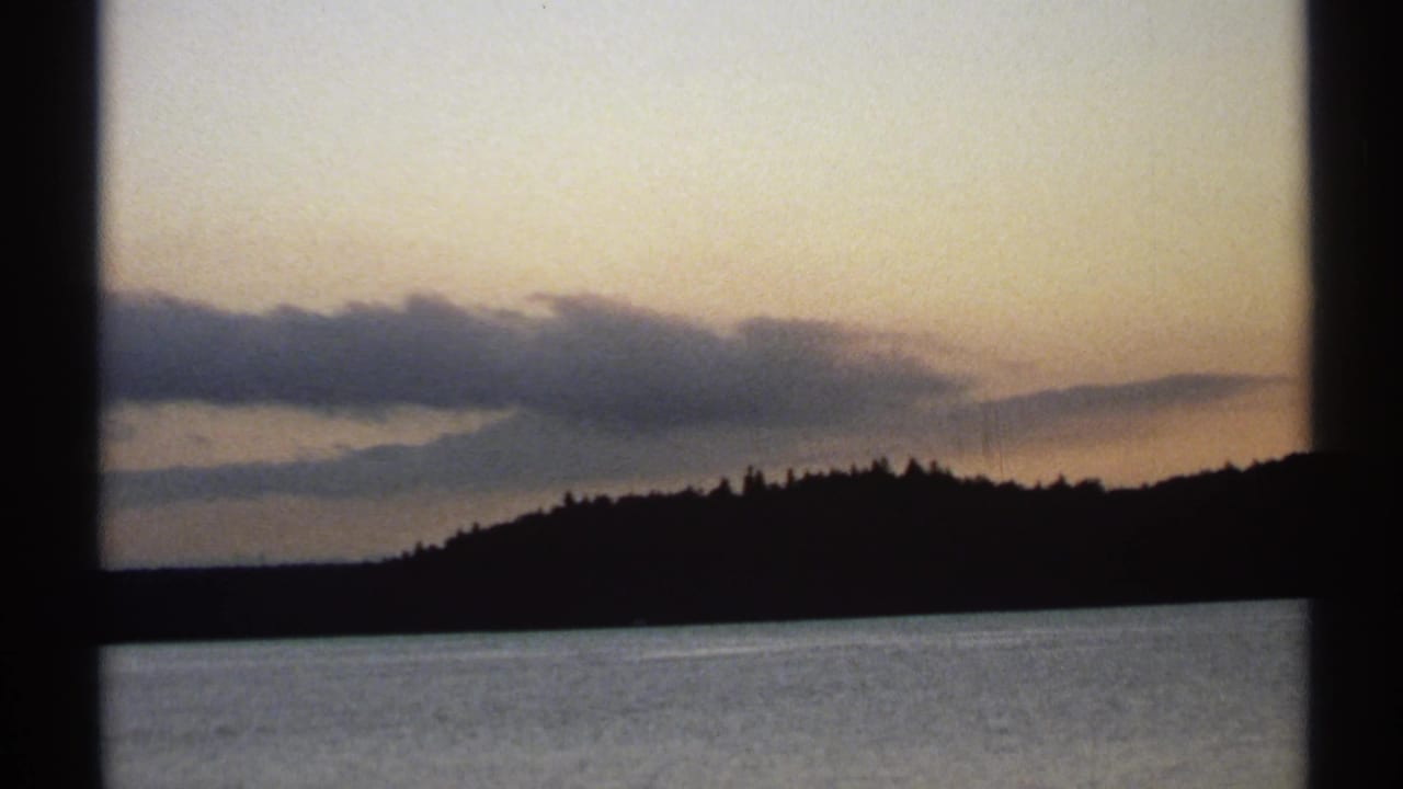 1971 New York 8mm frame showing a quiet lake, black tree-covered ridge, and muted evening clouds near the horizon.