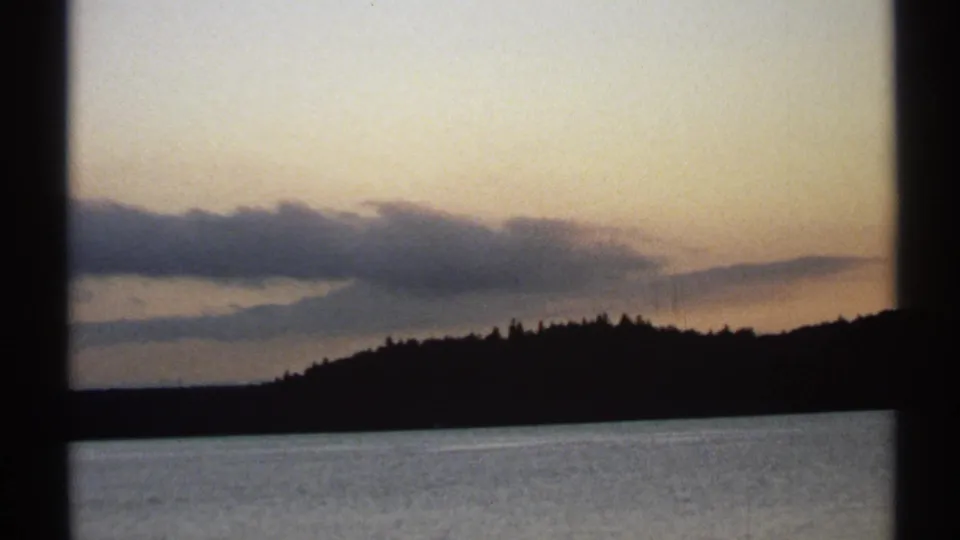 1971 New York 8mm frame showing a quiet lake, black tree-covered ridge, and muted evening clouds near the horizon.