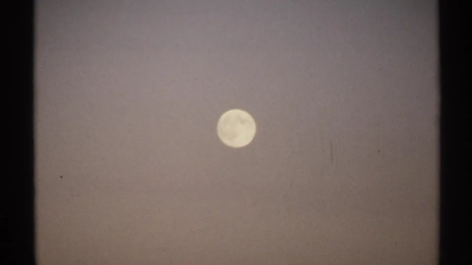 1971 8mm New York home-movie frame showing a pale full moon in a muted, hazy night sky with visible film edges.