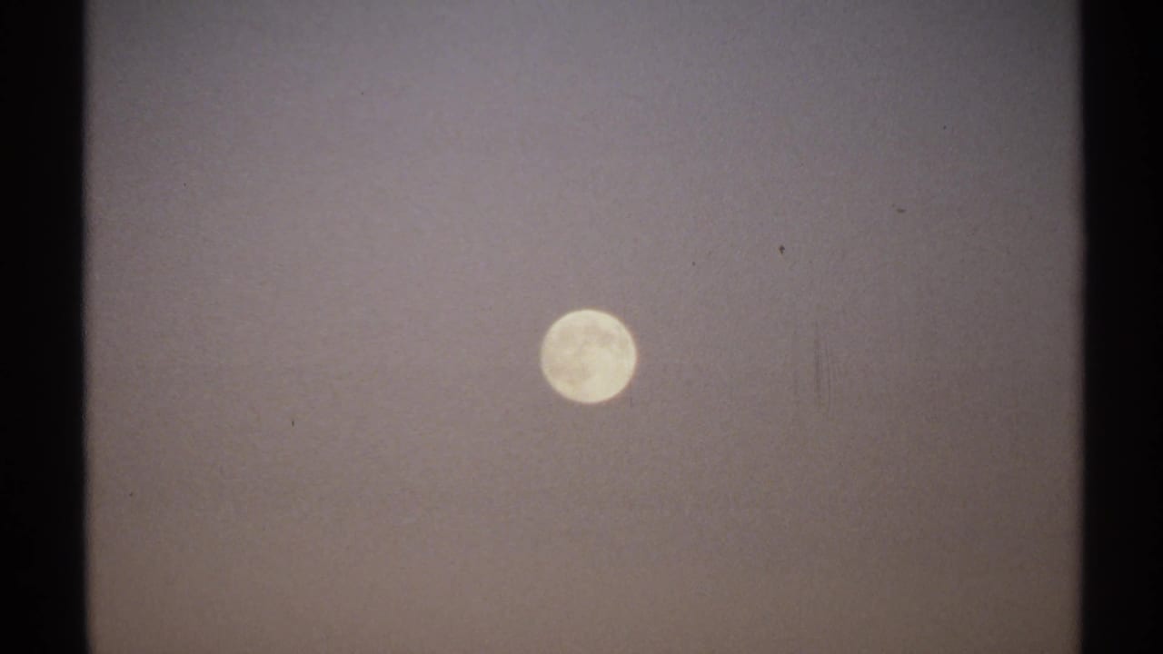 1971 8mm New York frame with the full moon held in a hazy sky, showing grain, slight softness, and contact-sheet cropping.