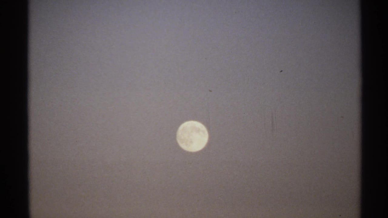 1971 8mm New York home-movie frame where the handheld camera places the full moon lower in a hazy, grainy sky.