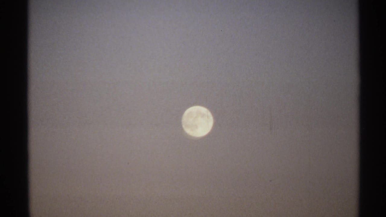 1971 8mm New York frame showing the full moon in a dim, hazy night sky with soft film grain and faint crater detail.