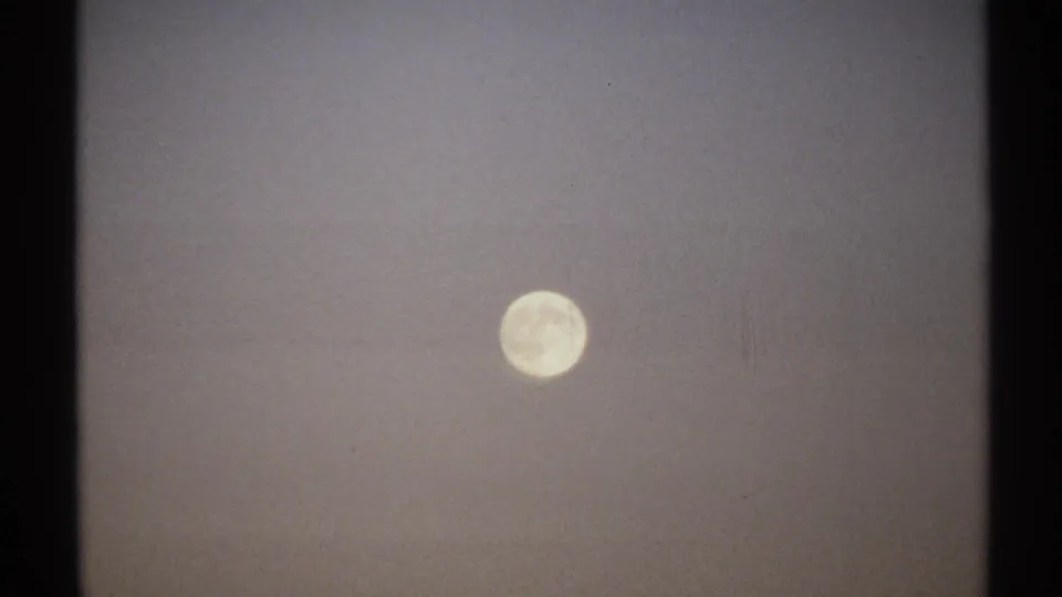 1971 8mm New York frame showing the full moon in a dim, hazy night sky with soft film grain and faint crater detail.