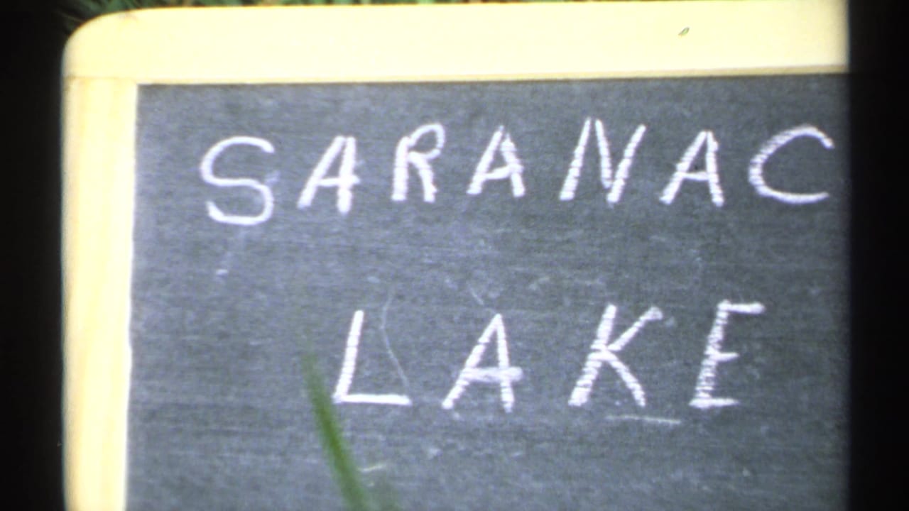1971 New York home-movie frame showing a dark chalkboard sign reading Saranac Lake, bordered by pale wood with slight film softness.