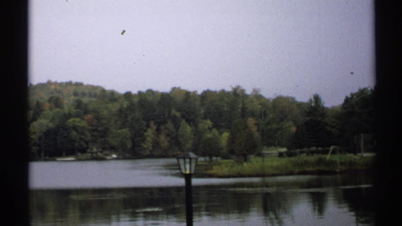 1971 New York lakeside view showing still water, wooded shore, low hills, pale sky, and a dark foreground lamppost.
