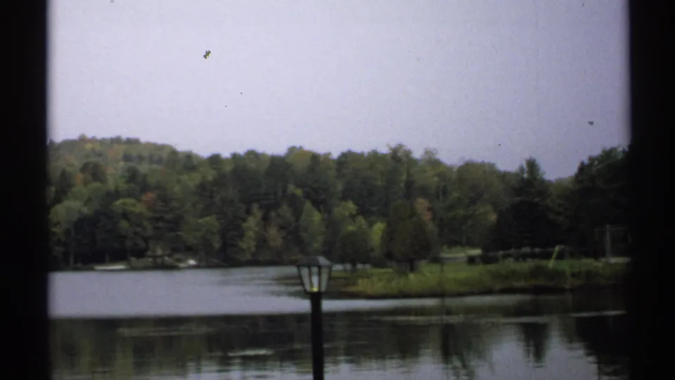 1971 New York lakeside view showing still water, wooded shore, low hills, pale sky, and a dark foreground lamppost.