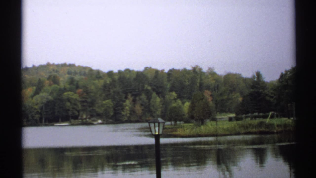 1971 New York 8mm frame of a tranquil Saranac Lake shoreline with wooded hills, reflective water, and a lamppost near the foreground.