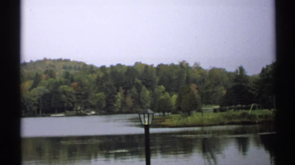 1971 New York 8mm frame of a tranquil Saranac Lake shoreline with wooded hills, reflective water, and a lamppost near the foreground.