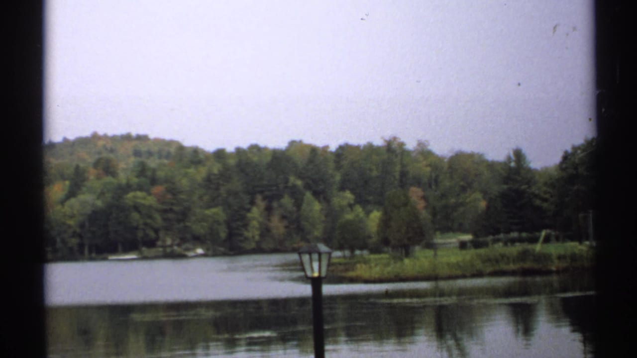 1971 New York 8mm frame showing calm lake water, wooded shoreline, early autumn foliage, and a dark foreground lamppost.
